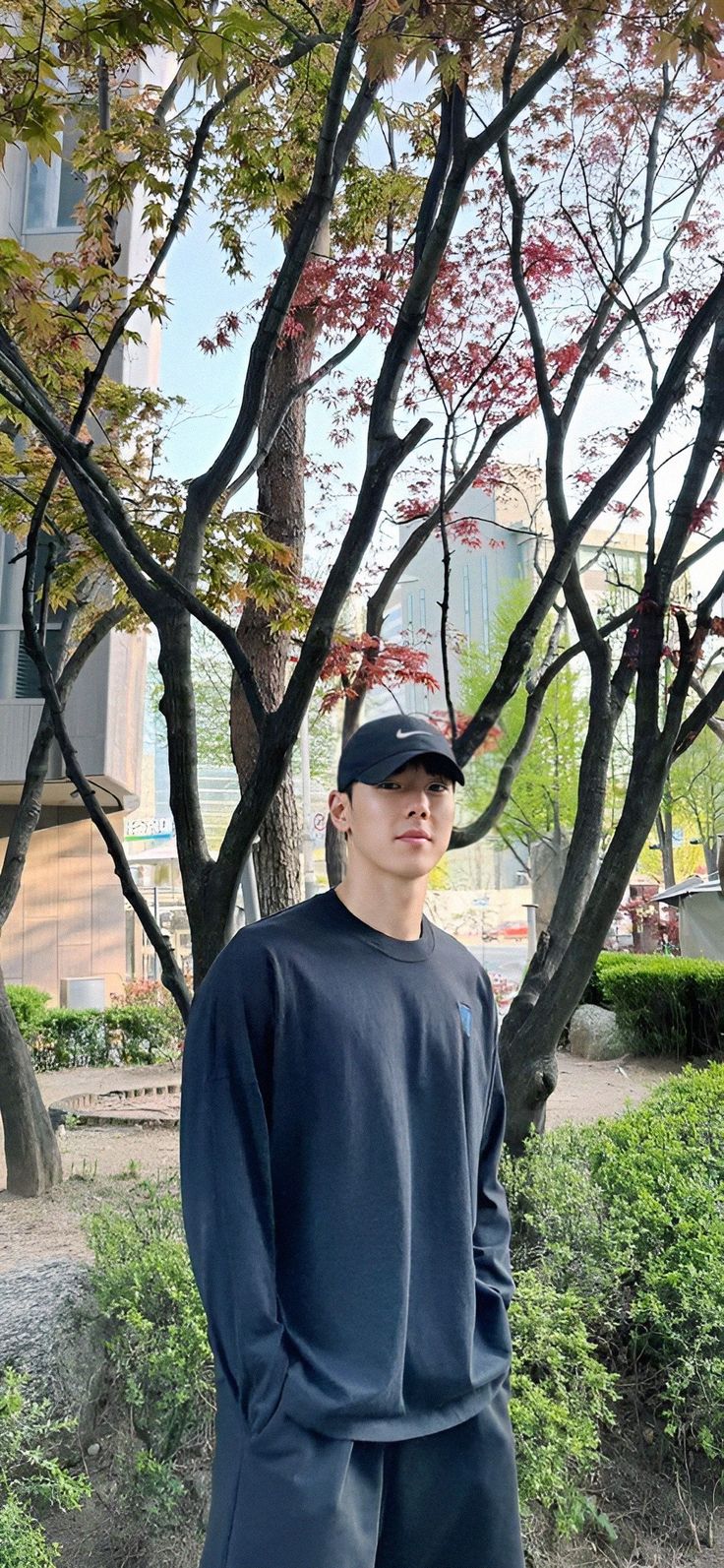 a man standing in front of a tree wearing a baseball cap and sweatpants with his hands on his hips