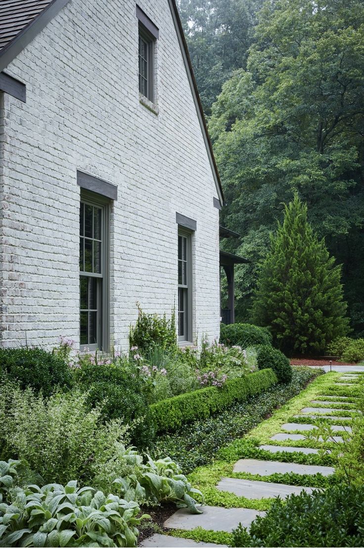 a white brick house surrounded by lush green plants and shrubs, with stone walkway leading to the front door