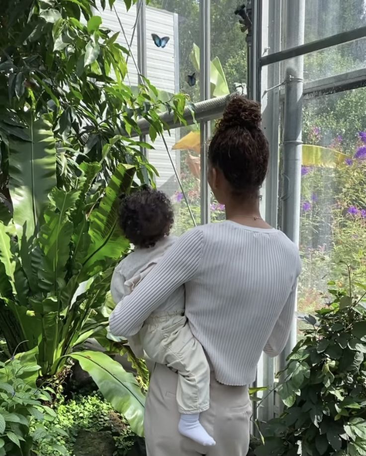 a woman holding a baby in her arms while looking at the plants inside of a greenhouse