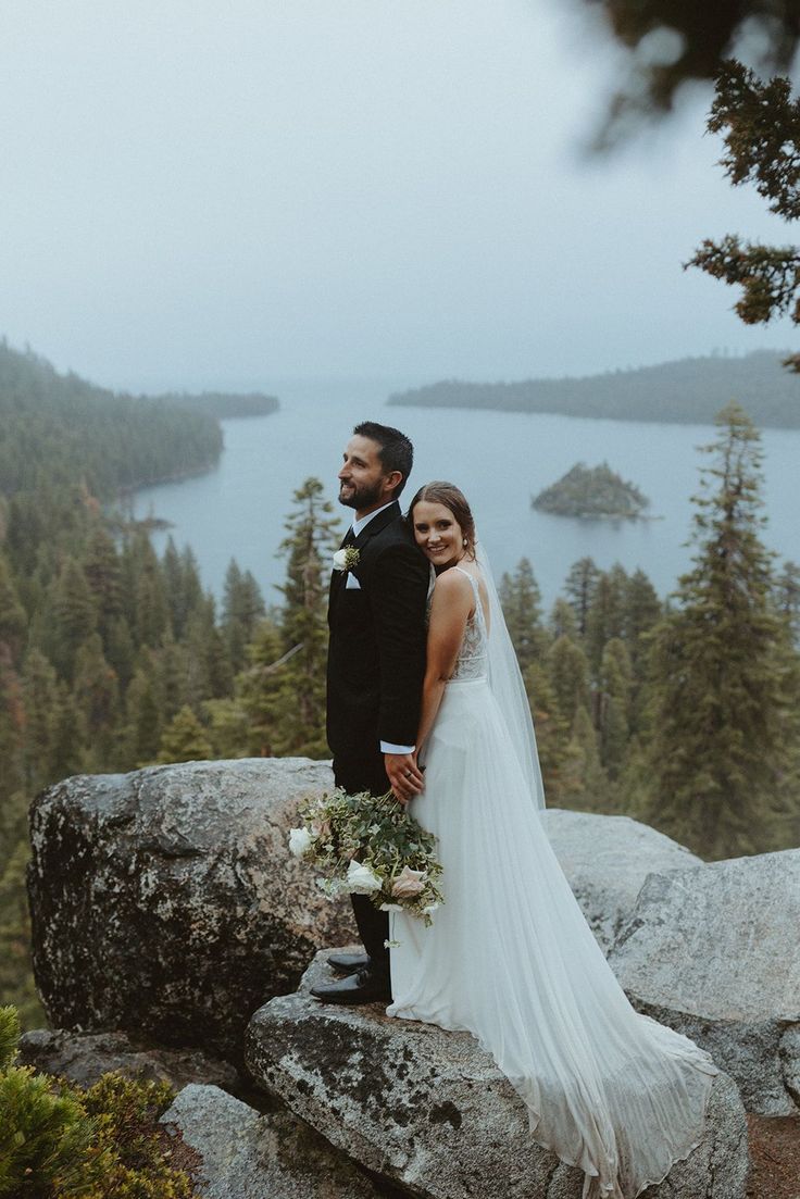 a bride and groom standing on top of a mountain looking at the camera with a lake in the background