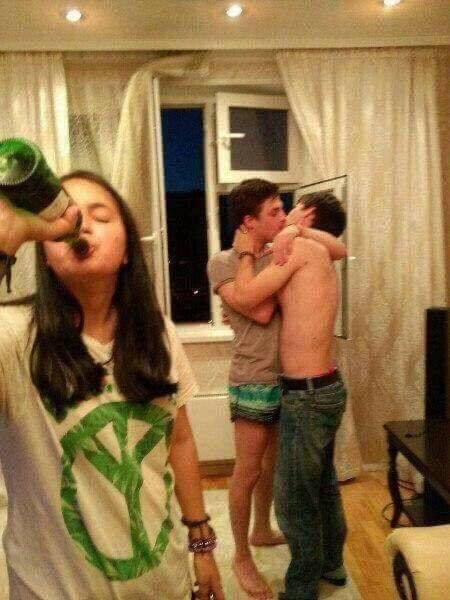 two men and a woman standing in a living room, one drinking from a bottle