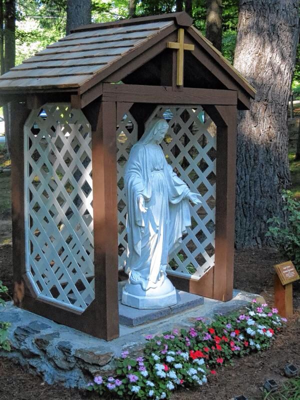 a statue in the middle of a garden with flowers around it and a wooden gazebo