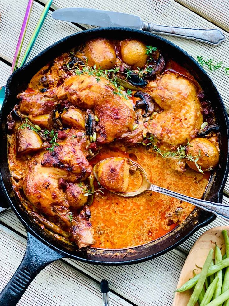 a skillet filled with chicken and potatoes on top of a wooden table next to green beans