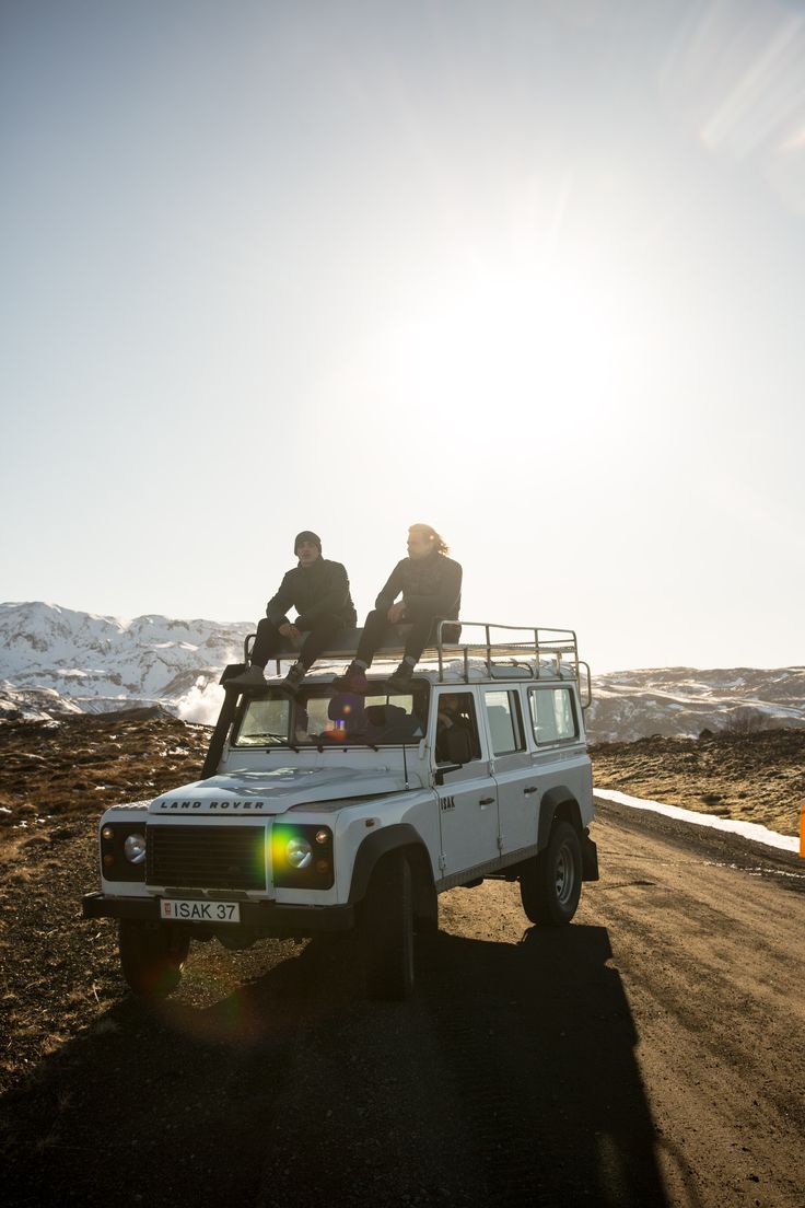 two people sitting on the roof of a white four door pick up truck driving down a dirt road