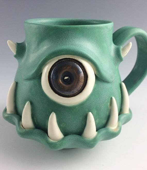 a green monster mug with white teeth and large eyes on it's side, sitting on a table