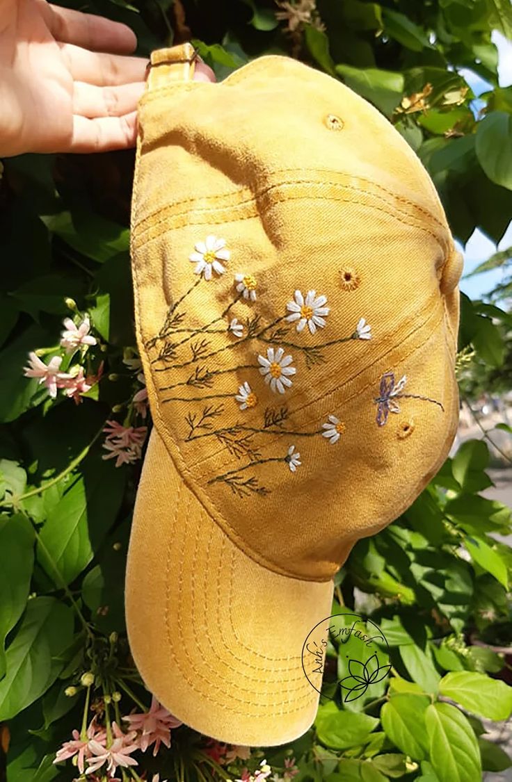 a yellow hat with daisies on it is hanging from a tree in front of some flowers