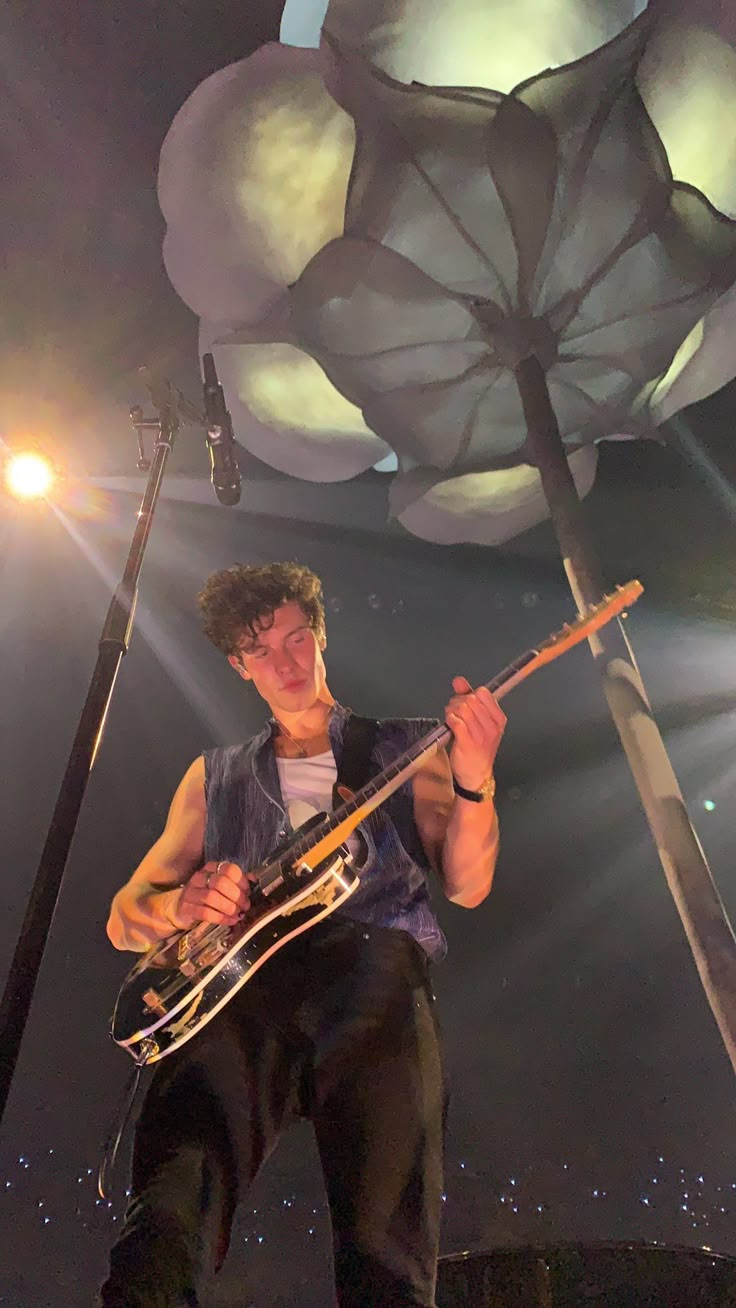 a man holding a guitar in front of a large light fixture on top of a stage