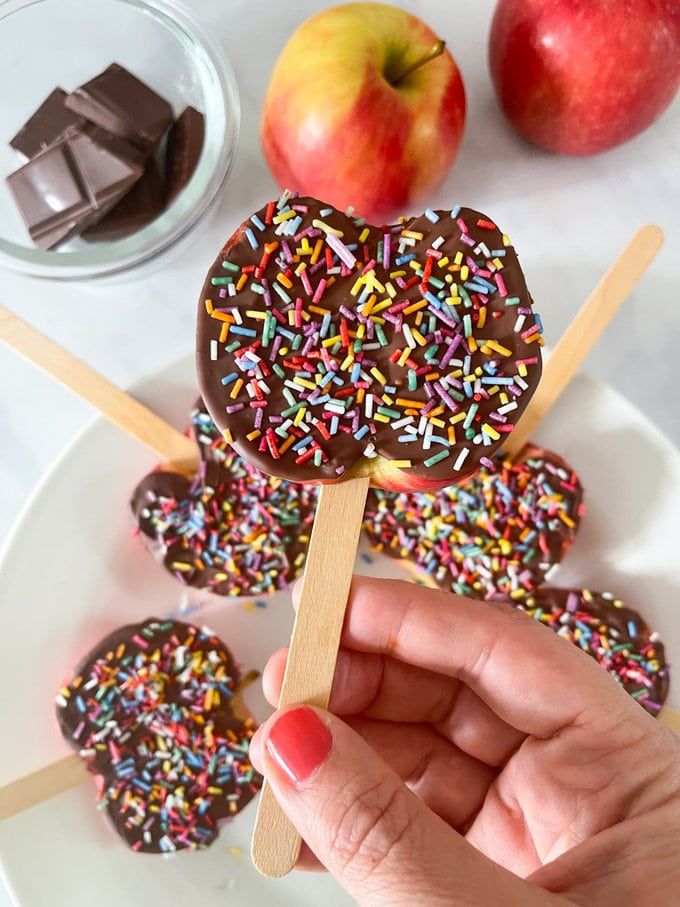 chocolate covered donuts with sprinkles on a stick and an apple in the background