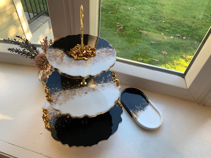 three tiered black and white cake with gold decorations on it sitting in front of a window