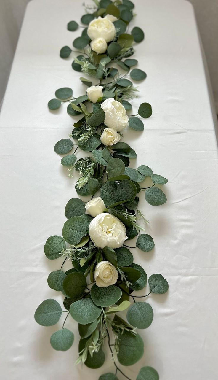 a long table with white flowers and greenery on it's sides, along with green leaves