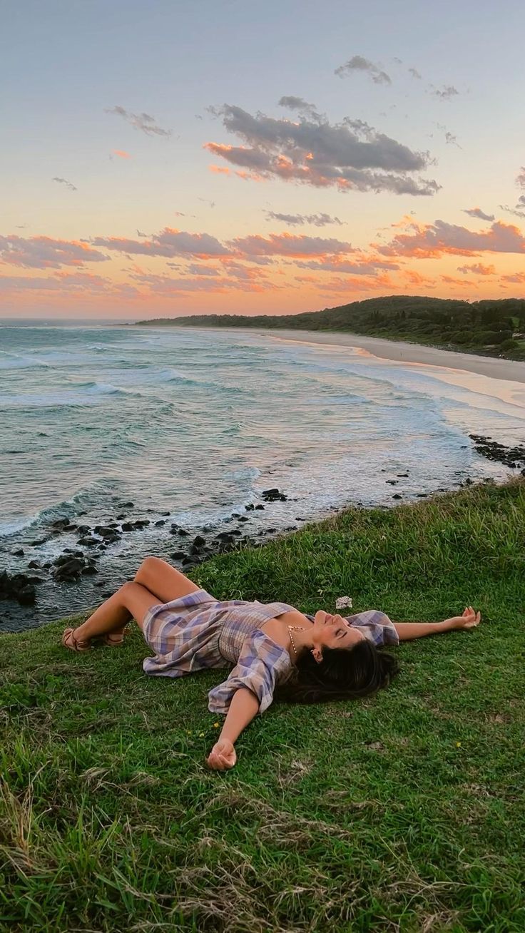 a woman laying on top of a lush green field next to the ocean at sunset