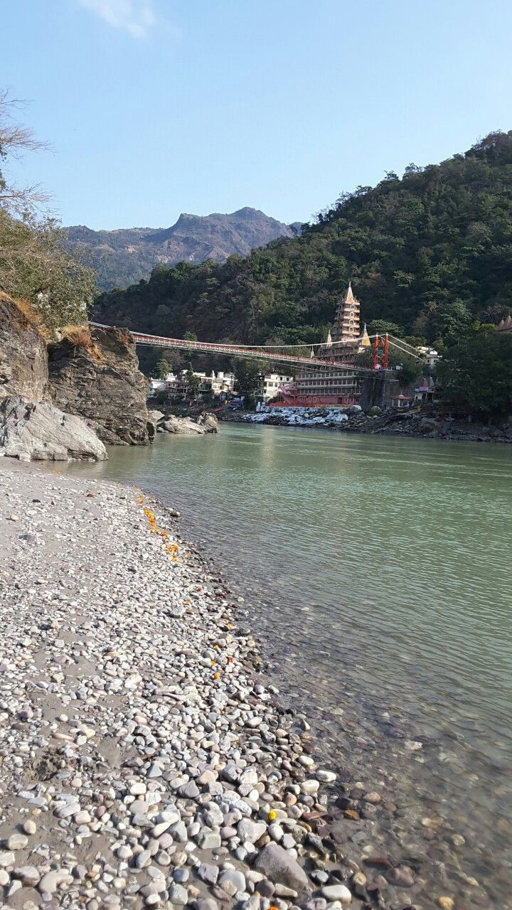 a river with rocks on the shore and a bridge in the background, surrounded by mountains