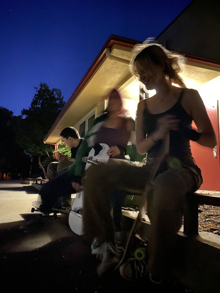 three people sitting outside at night with one person on a skateboard and the other standing