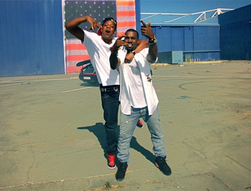 two men standing in an empty parking lot with their arms up and one pointing at the camera