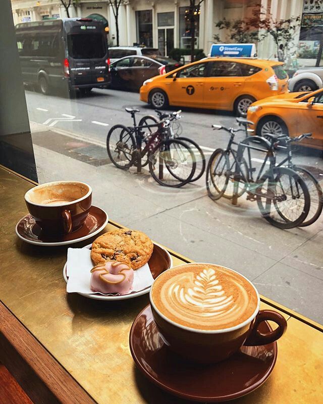 two cups of coffee sit on a table with pastries and doughnuts in front of a window