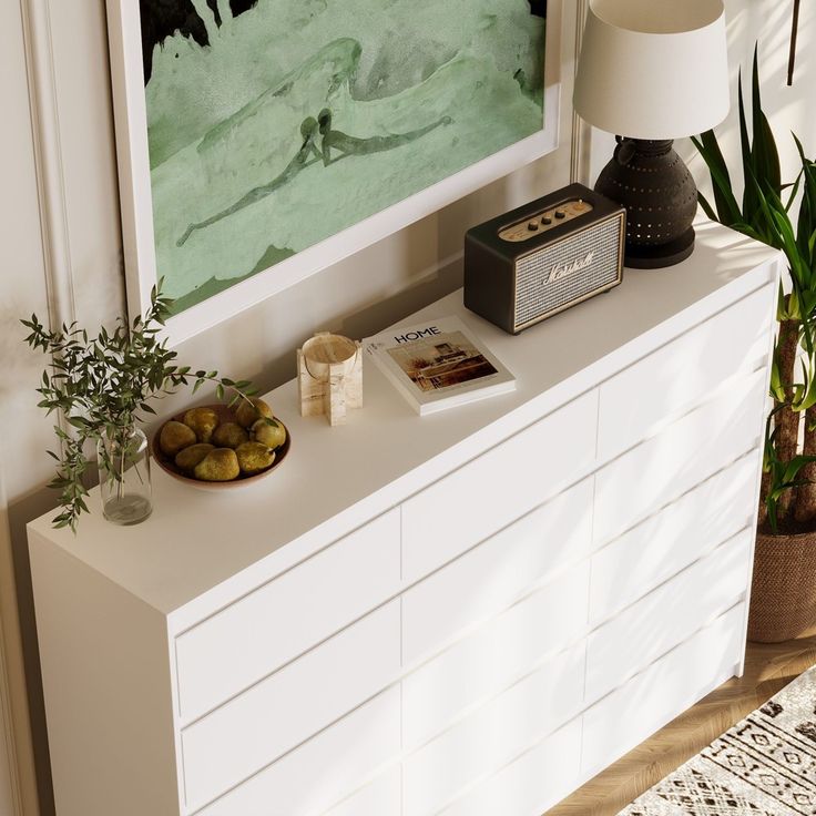 a white dresser topped with a bowl of fruit next to a painting