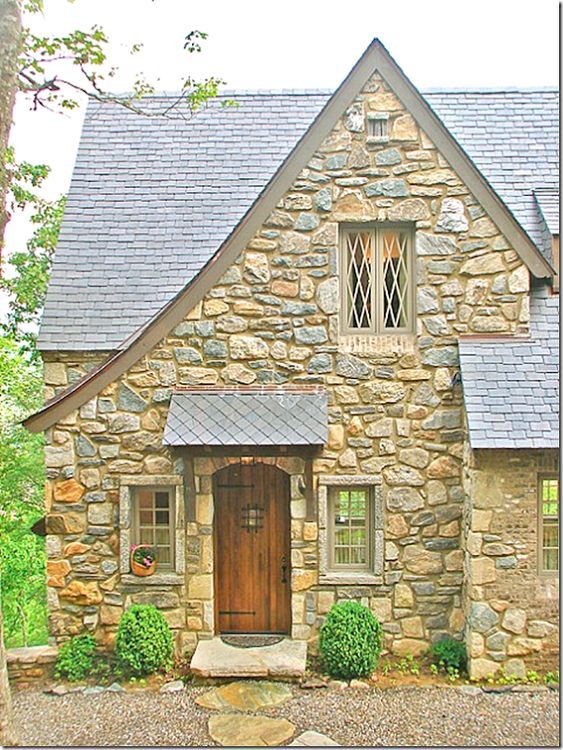 a stone house with a wooden door and window on the front porch is surrounded by greenery