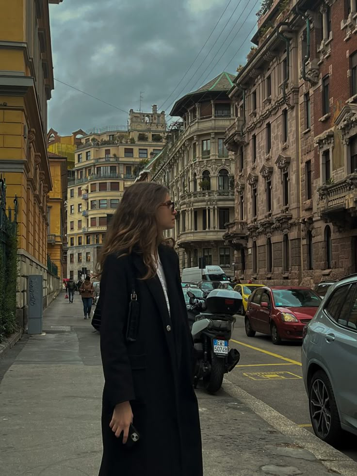 a woman standing on the side of a street next to parked cars and tall buildings