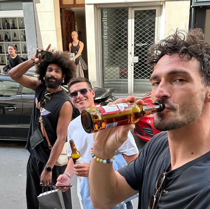 a man is drinking from a beer bottle while others are standing on the street behind him