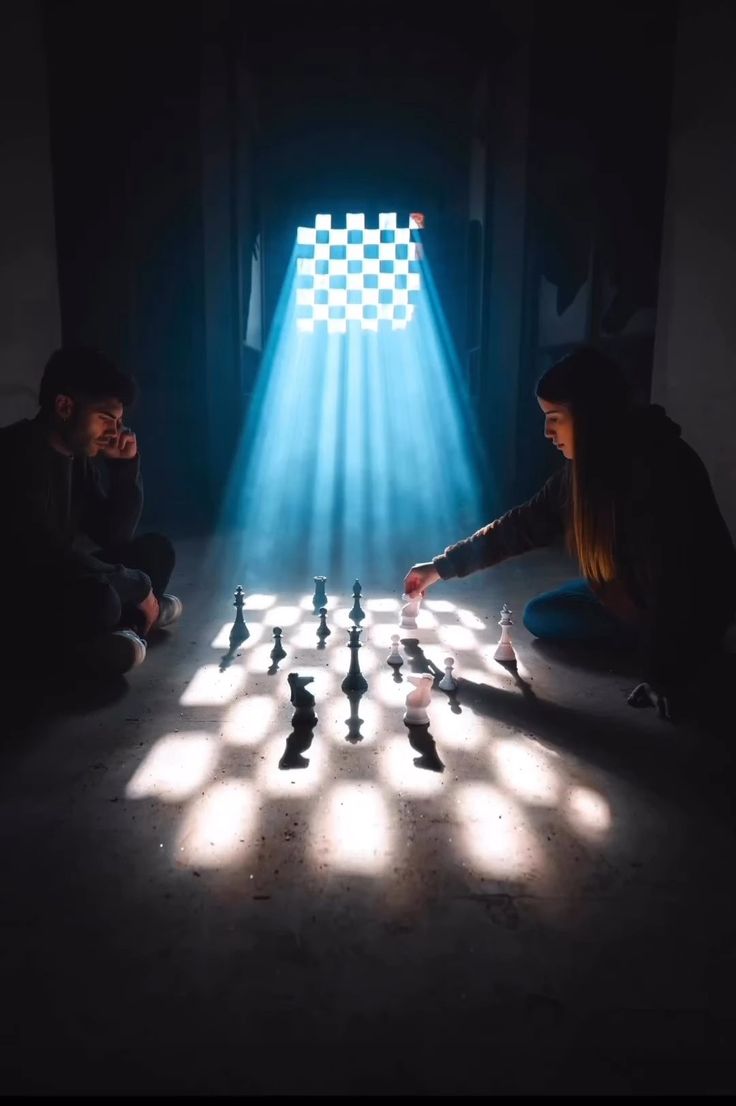 two people sitting on the floor playing chess in front of a light that is shining down on them