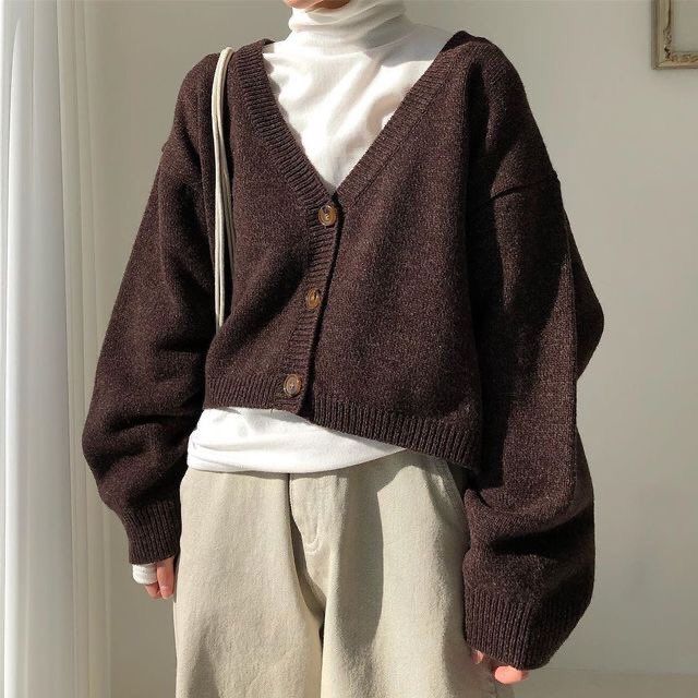 a woman standing in front of a white wall wearing a brown sweater