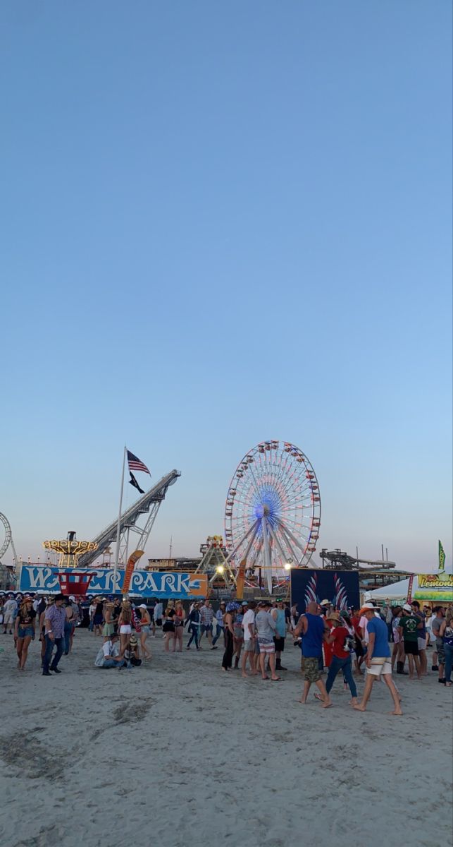 many people are walking on the beach near carnival rides and ferris wheel in the distance