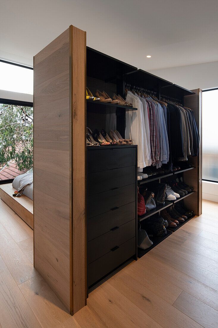 an open closet with clothes and shoes on the shelves next to a bed in front of large windows