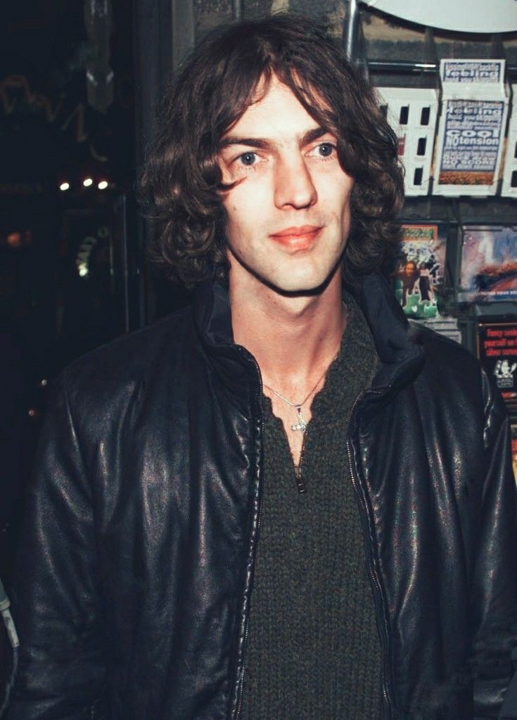 a man with long hair wearing a black leather jacket and looking at the camera while standing in front of a store