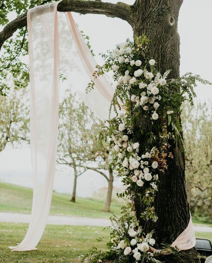 an outdoor ceremony setup with white flowers and greenery on the tree trunk, along with sheer draping