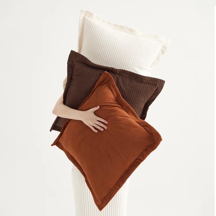 a woman is holding three pillows in her hands, one brown and the other white