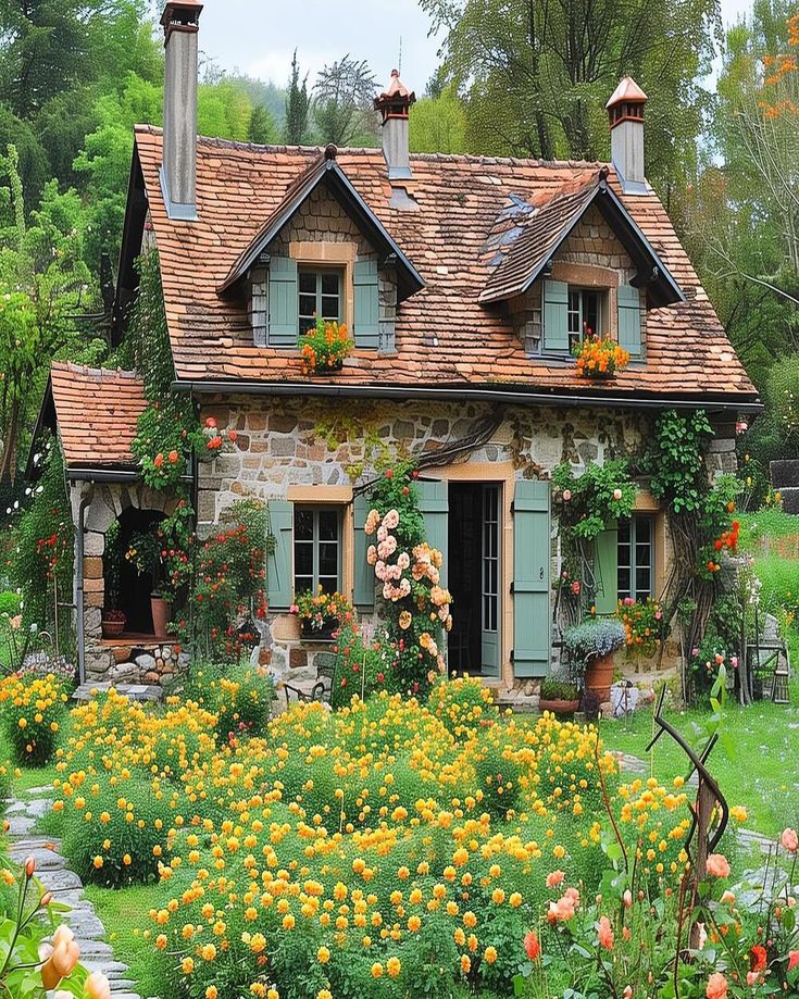 an old stone house with flowers in the front yard and windows on each side, surrounded by greenery