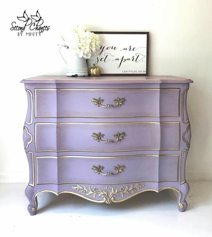 a purple dresser sitting next to a white vase with flowers on top and a sign above it