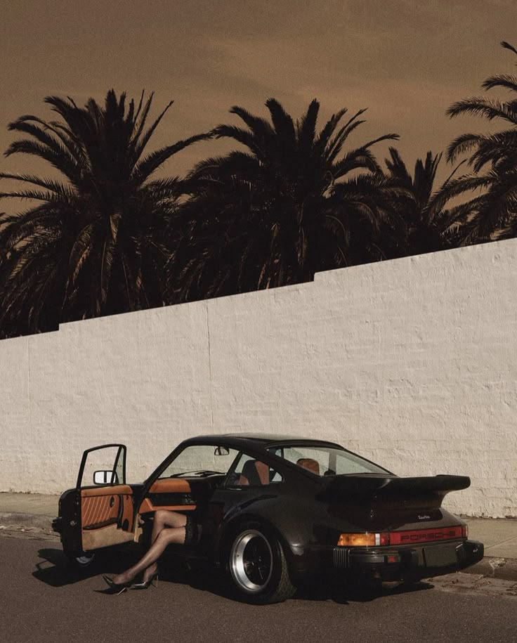 a woman sitting in the back of a car next to a white wall and palm trees