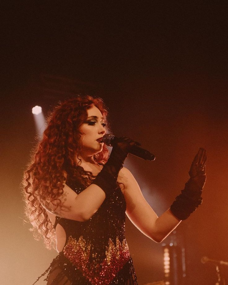 a woman with long red hair and black gloves on her hands is singing into a microphone