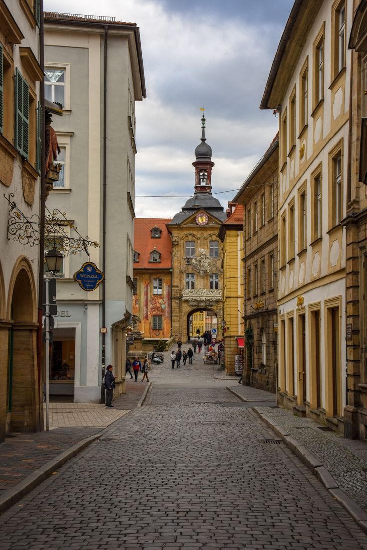 people are walking down an old cobblestone street with buildings on both sides and a clock tower in the distance