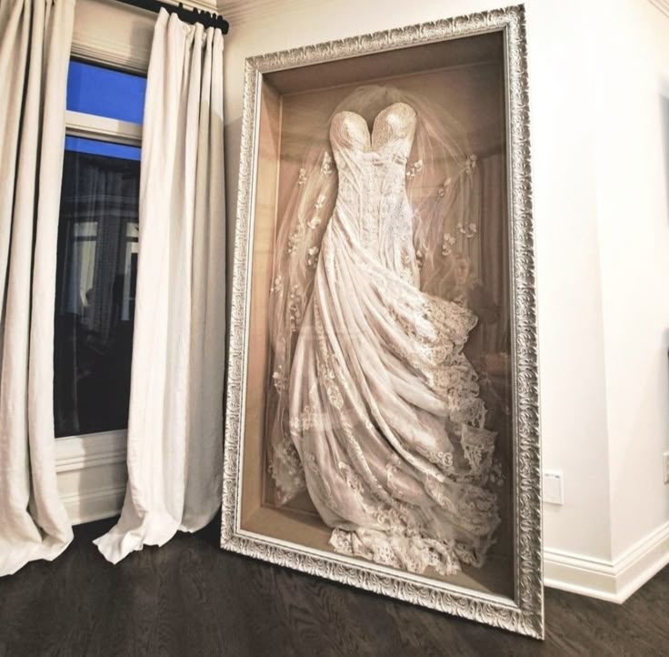 a wedding dress is displayed in an ornate frame on the floor next to a window