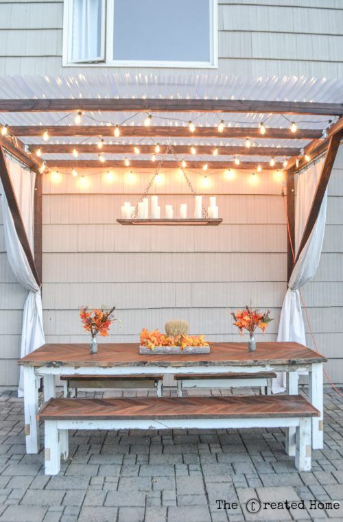 an outdoor dining area with a table and bench under the pergolated canopy that has lights strung over it