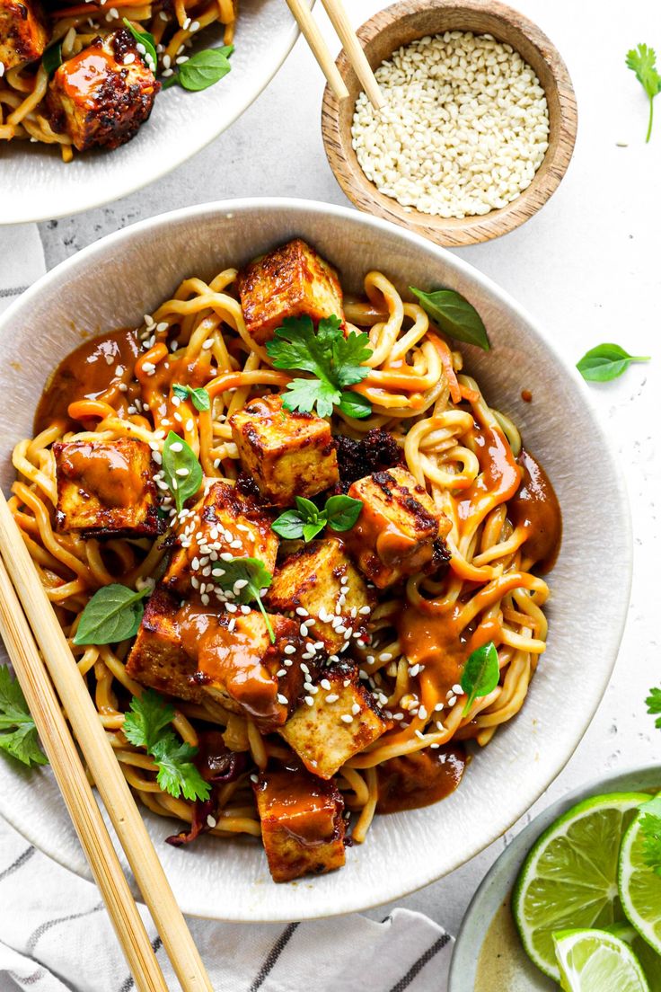 two bowls filled with noodles and tofu on top of a white table next to chopsticks