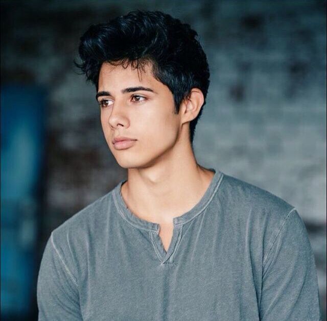 a young man with black hair is standing in front of a brick wall and looking at the camera