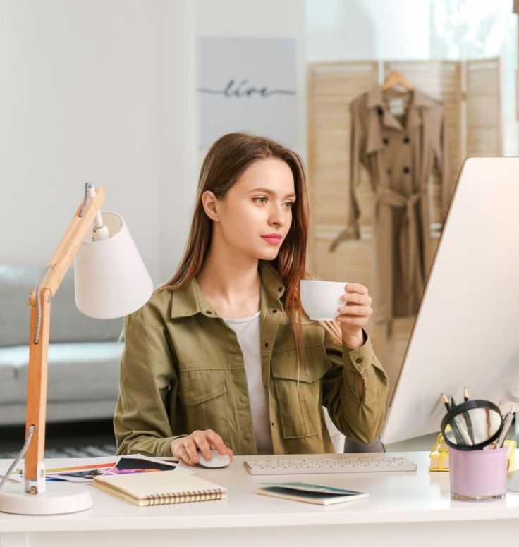 a woman sitting at a desk with a cup in her hand and looking at the computer screen