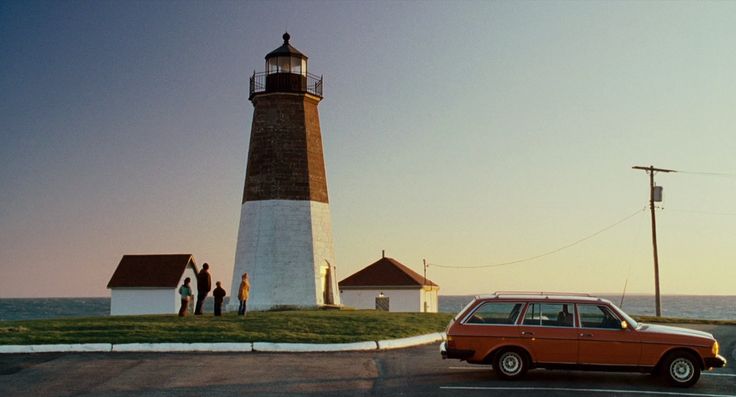 an orange car parked in front of a light house with people standing on the side