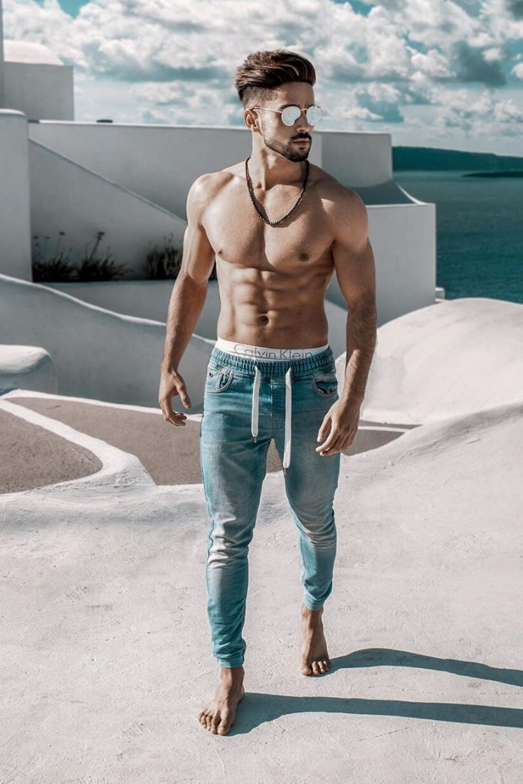 a man with no shirt standing in front of the ocean wearing blue jeans and an eye patch