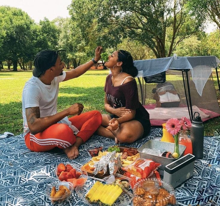 a man and woman are sitting on a blanket in the park eating fruit from a cooler