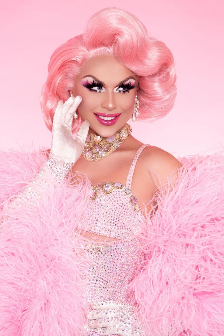 a woman with pink hair and makeup is wearing a pink feathered coat, holding her hand up to her face