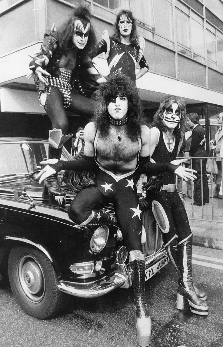 the kiss band posing on top of a car in front of a building with people standing around