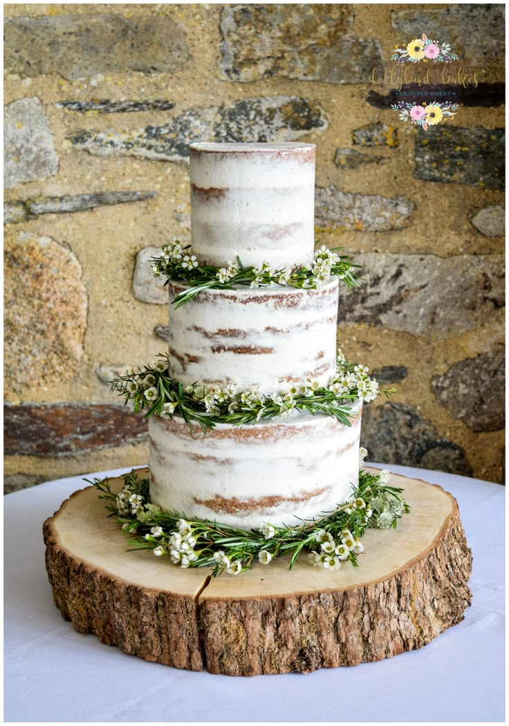 a three tiered wedding cake with greenery on top sits on a tree stump