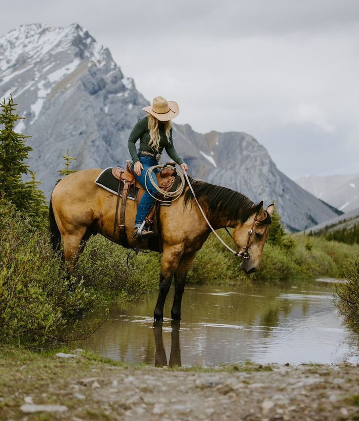 a woman riding on the back of a brown horse next to a river and mountains