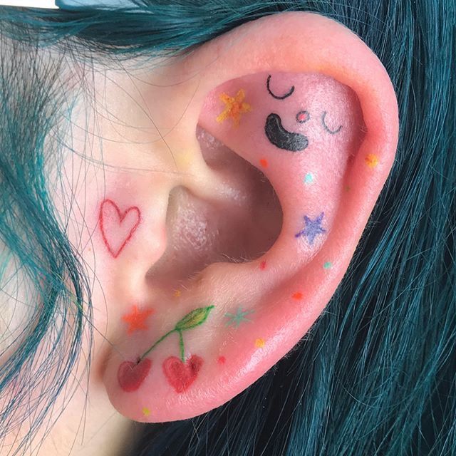 a girl with blue hair has colorful tattoos on her ear and behind the ear are stars, hearts, and clouds