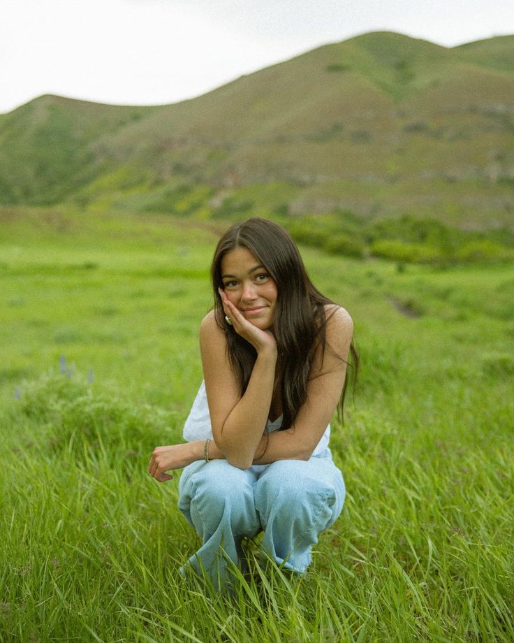 a woman sitting in the grass with her hand on her chin and looking at the camera