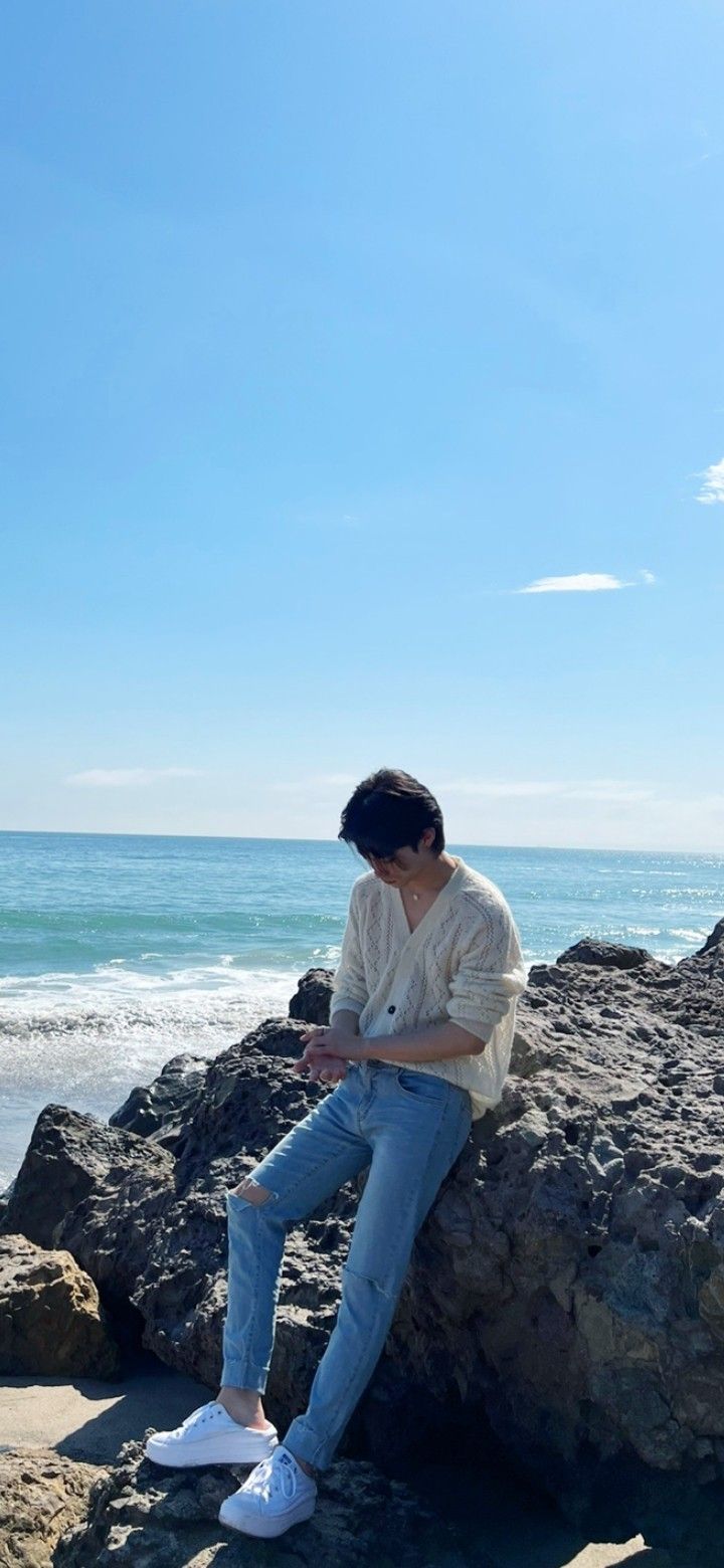 a person sitting on rocks by the ocean looking at their cell phone while wearing white sneakers